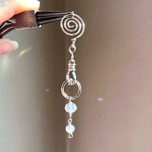 Puede incluir: Pendiente plateado hecho a mano con una espiral, un pequeño dije de mano y dos cuentas de cristal iridiscente. El pendiente se sostiene con unos alicates sobre un fondo suave y neutro.