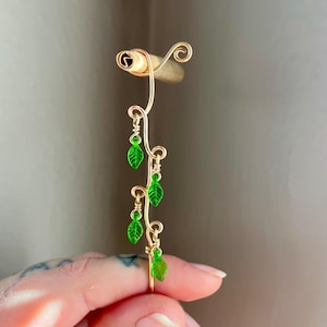 May include: A gold-coloured wire ring with a vine design. The vine features small green leaf charms. The ring is held by a hand against a neutral background. The ring's design is delicate and nature-inspired.
