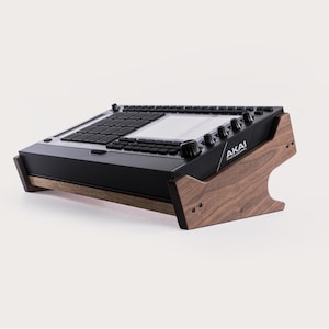 May include: A black Akai Professional music production center on a wooden stand. The stand is made of dark brown wood and has a curved design. The music production center has a screen, buttons, and knobs. The Akai logo is visible on the side.