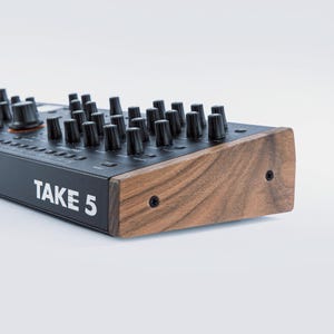 May include: Close-up of a black synthesizer with numerous black knobs and the text "TAKE 5" in white. The side panel is made of wood, and the instrument has a modern design.
