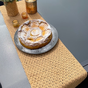 May include: A brown crocheted table runner with a white cake topped with orange slices and powdered sugar on a black plate on a grey table.