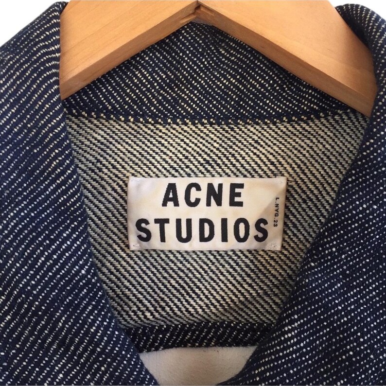 Puede incluir: Primer plano de una chaqueta vaquera azul con una etiqueta blanca que dice "Acne Studios" en letras negras.