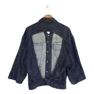 Puede incluir: Una chaqueta vaquera azul oscuro con un dise&ntilde;o &uacute;nico que presenta un corte en la espalda, revelando un panel de mezclilla contrastante. La chaqueta tiene un cierre de botones en la parte delantera y mangas largas.