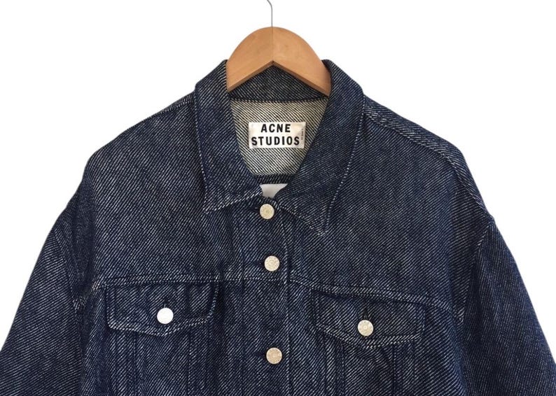 Puede incluir: Una chaqueta vaquera azul oscuro con cierre de botones y una etiqueta blanca que dice "Acne Studios".