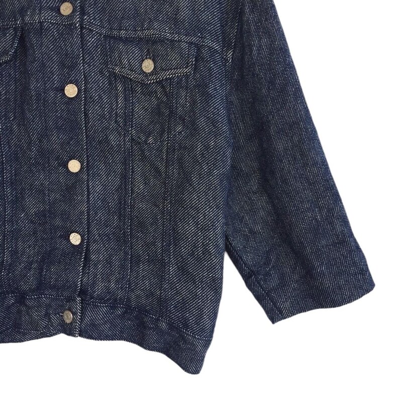 Puede incluir: Una chaqueta vaquera azul oscuro con cierre de botones y un solo bolsillo en el pecho. La chaqueta tiene un corte ligeramente holgado y una tela texturizada.