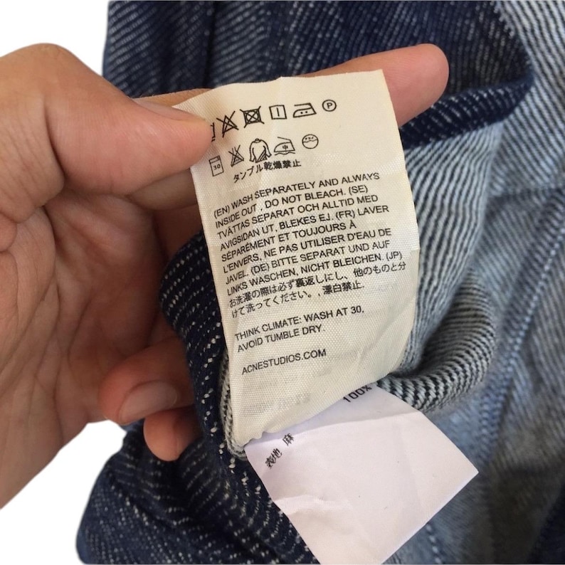 Puede incluir: Una etiqueta de cuidado blanca con texto negro que detalla las instrucciones de lavado para una prenda de vestir. El texto incluye instrucciones en ingl&eacute;s, sueco, franc&eacute;s, alem&aacute;n y japon&eacute;s. La etiqueta tambi&eacute;n incluye un gr&aacute;fico de una lavadora, una plancha y un s&iacute;mbolo de limpieza en seco. El texto dice "Think Climate: Wash at 30, Avoid Tumble Dry. ACNESTUDIOS.COM"