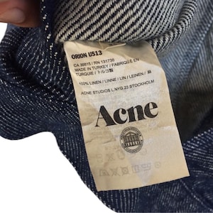 Puede incluir: Una camisa de lino a rayas azul y blanca con una etiqueta blanca que dice "ORION US13", "CA 39815/RN 131738", "MADE IN TURKEY/FABRIQU&Eacute; EN TURQUIE/トルコ製", "100% LINEN/LINNE/LIN/LEINEN/麻", "ACNE STUDIOS L.NYG.23 STOCKHOLM" y "Acne".