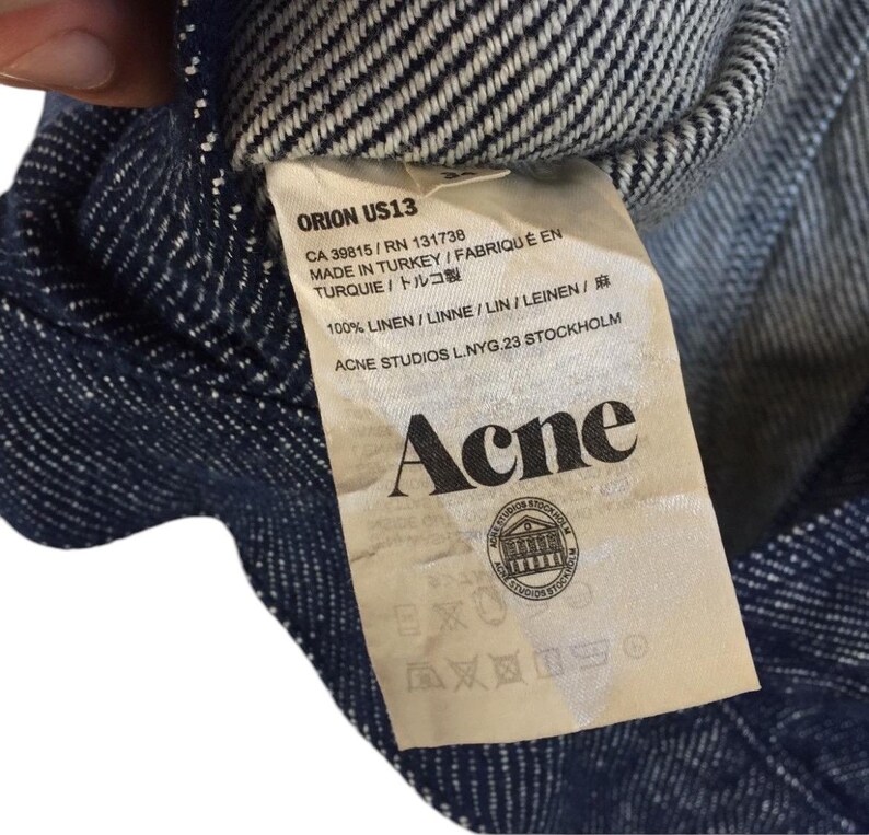 Puede incluir: Una camisa de lino a rayas azul y blanca con una etiqueta blanca que dice "ORION US13", "CA 39815/RN 131738", "MADE IN TURKEY/FABRIQU&Eacute; EN TURQUIE/トルコ製", "100% LINEN/LINNE/LIN/LEINEN/麻", "ACNE STUDIOS L.NYG.23 STOCKHOLM" y "Acne".