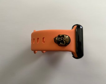 May include: A peach-colored silicone watch band with a black watch face and a gold skull charm.