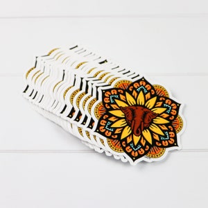 May include: A stack of stickers featuring a colorful mandala design with an elephant's head in the center. The mandala is in shades of orange, yellow, and blue, with a black outline. The elephant's head is brown with white tusks.
