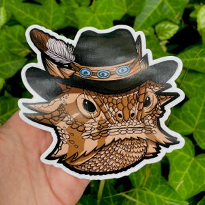 K&ouml;nnte beinhalten: Eine Cartoon-Illustration eines Bartagamen, der einen schwarzen Cowboyhut mit einer braunen Feder und einem Perlenband tr&auml;gt. Der Drache hat einen braunen und orangefarbenen schuppigen K&ouml;rper und einen ernsten Ausdruck.