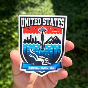 K&ouml;nnte beinhalten: Ein Aufkleber mit einem blauen, wei&szlig;en und roten Schild mit dem Text "UNITED STATES NATIONAL DIVING TEAM". Das Schild zeigt einen Taucher im Wasser mit dem Space Needle in Seattle im Hintergrund.