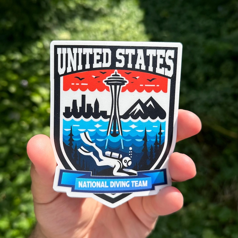 K&ouml;nnte beinhalten: Ein Aufkleber mit einem blauen, wei&szlig;en und roten Schild mit dem Text "UNITED STATES NATIONAL DIVING TEAM". Das Schild zeigt einen Taucher im Wasser mit dem Space Needle in Seattle im Hintergrund.