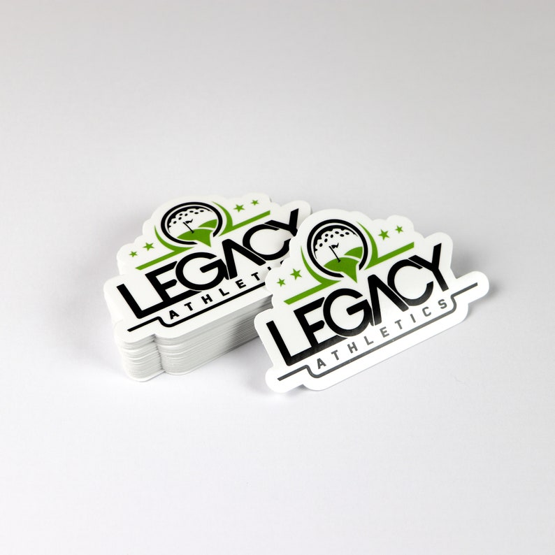 May include: A stack of white, round stickers with a black and green logo that says "LEGACY ATHLETICS". The logo features a golf ball with a flag on a green field.