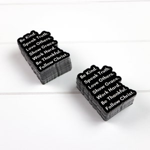 May include: A stack of black square stickers with white text that reads "Be Kind. Speak Truth. Love Others. Show Grace. Work Hard. Be Thankful. Follow Christ."