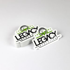 K&ouml;nnte beinhalten: Ein Stapel wei&szlig;er, runder Aufkleber mit einem schwarzen und gr&uuml;nen Logo, auf dem "LEGACY ATHLETICS" steht. Das Logo zeigt einen Golfball mit einer Fahne darauf.