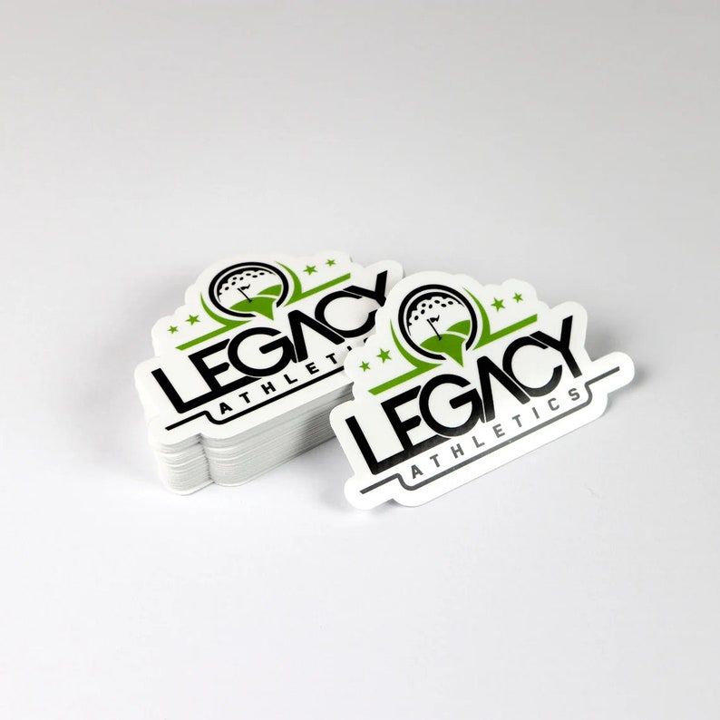 K&ouml;nnte beinhalten: Ein Stapel wei&szlig;er, runder Aufkleber mit einem schwarzen und gr&uuml;nen Logo, auf dem "LEGACY ATHLETICS" steht. Das Logo zeigt einen Golfball mit einer Fahne darauf.