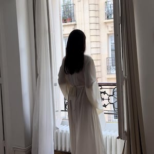 May include: A floor-length off-white silk robe with wide sleeves and a self-tie belt. The robe is shown hanging loosely on a person standing in front of a window overlooking a Parisian street. The robe is made of a lightweight, flowing fabric, and the overall aesthetic is minimalist and elegant.