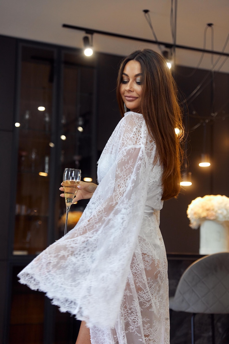 May include: A white lace robe with long sleeves. The robe is worn with a matching white outfit and a glass of champagne. The background is a dark room with soft lighting.