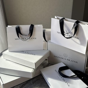 May include: Several white shopping bags and boxes with black ribbon handles are stacked on a gray surface. The bags and boxes are labeled 'MONIKA BLANK ATELIER' in a minimalist black sans-serif font.  The packaging is uniform in style, suggesting a cohesive brand identity. The image showcases luxury packaging for retail or e-commerce purposes.