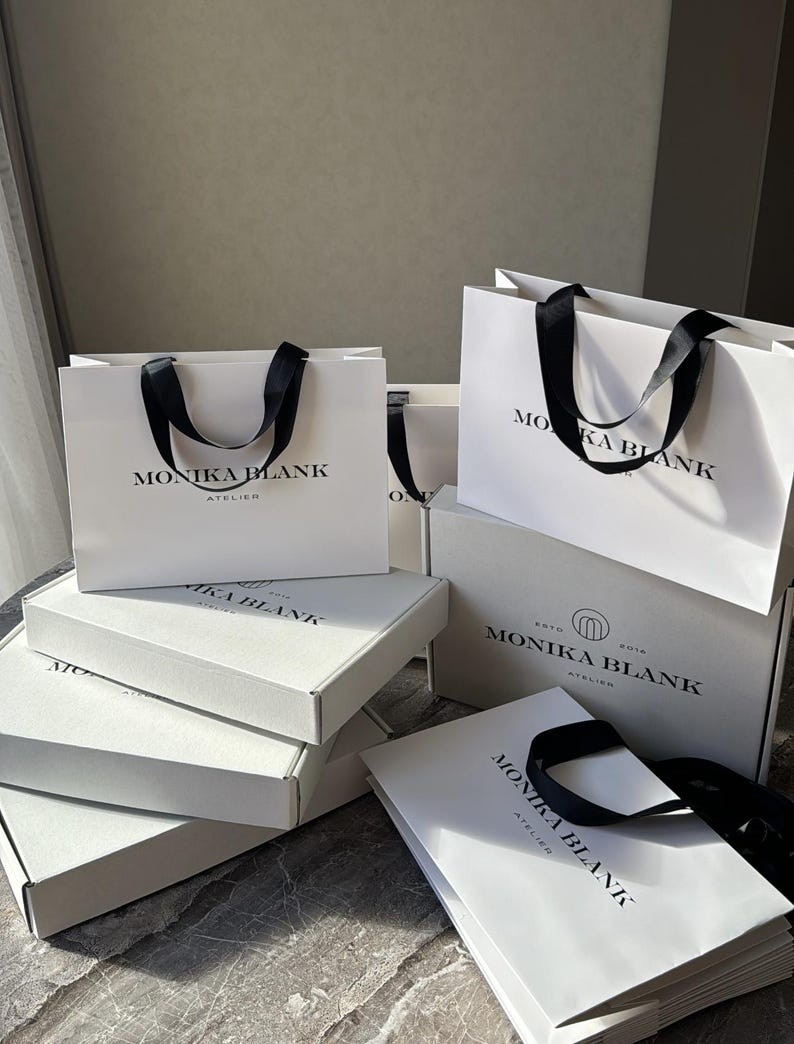 May include: Several white shopping bags and boxes with black ribbon handles are stacked on a gray surface. The bags and boxes are labeled 'MONIKA BLANK ATELIER' in a minimalist black sans-serif font.  The packaging is uniform in style, suggesting a cohesive brand identity. The image showcases luxury packaging for retail or e-commerce purposes.