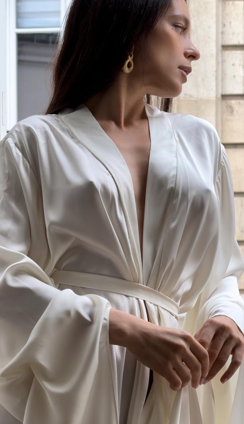 May include: A luxurious off-white satin robe with long sleeves and a self-tie belt. The robe features a relaxed fit and a deep V-neckline. The fabric drapes elegantly, creating soft folds and creases. The robe is shown against a blurred background, suggesting a relaxed and comfortable atmosphere.  The overall aesthetic is sophisticated and minimalist.