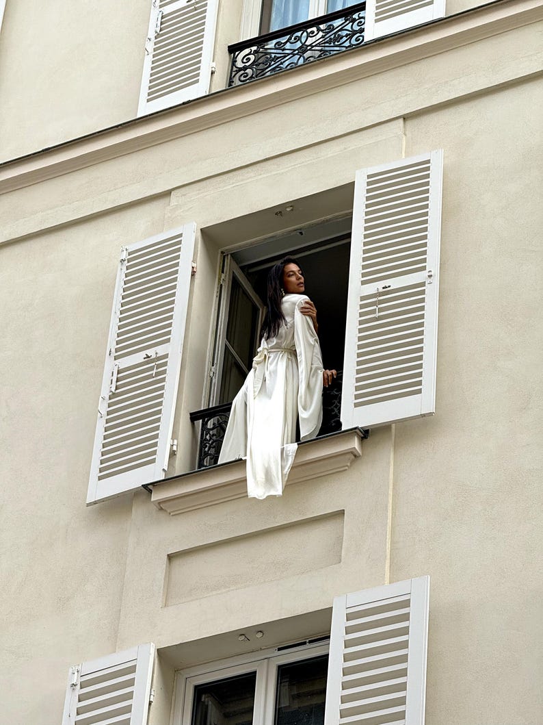 May include: A woman stands on a balcony, wearing a flowing, light beige silk robe. The robe has long sleeves and falls to her ankles. She is framed by a window with white shutters, set in a light beige building. The balcony has a simple black metal railing. The overall aesthetic is minimalist and elegant.