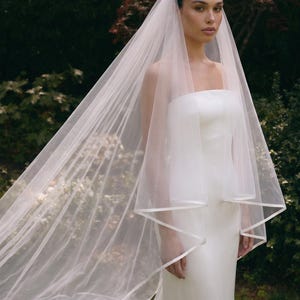 May include: A long, sheer white bridal veil featuring a delicate satin ribbon trim along the edges. The veil is draped over a strapless white gown, extending past the shoulders and flowing down to create a soft, layered appearance.