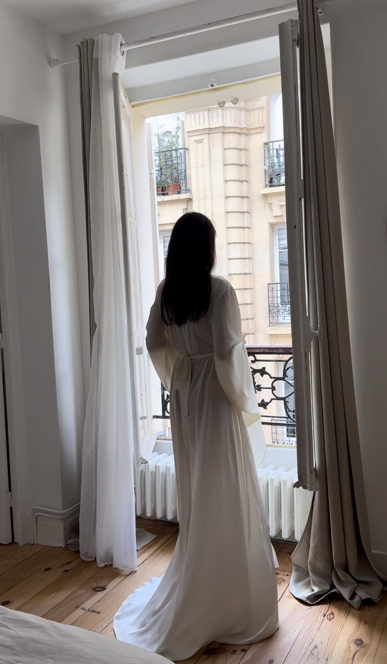 May include: A floor-length off-white silk robe with wide sleeves and a self-tie belt. The robe is shown in a room with hardwood floors and sheer white curtains framing a window with a view of a Parisian street.