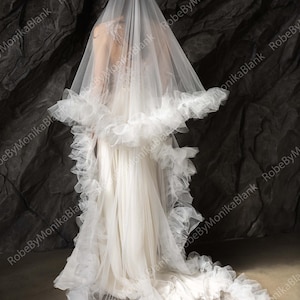 May include: A long, sheer white bridal veil featuring a voluminous, ruffled tulle trim along the edges. The veil is draped over the back, extending down to the floor in a train, with a delicate, layered texture and a soft, translucent appearance.