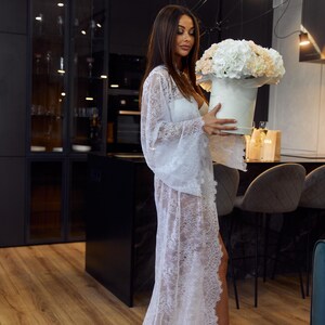 May include: A white lace robe with bell sleeves and a high slit. The robe is worn over a white outfit. A round white box holds a bouquet of white and peach flowers. The setting is a modern kitchen with dark cabinets and wood floors.