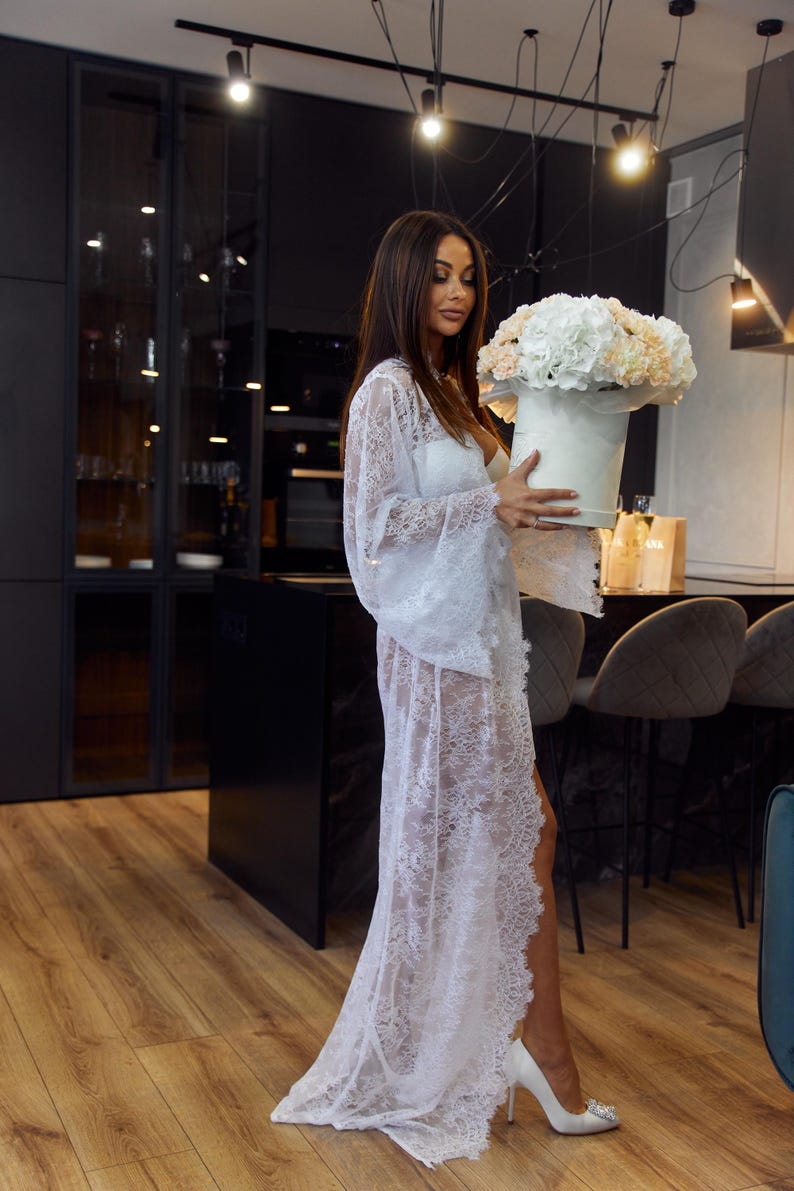 May include: A white lace robe with bell sleeves and a high slit. The robe is worn over a white outfit. A round white box holds a bouquet of white and peach flowers. The setting is a modern kitchen with dark cabinets and wood floors.