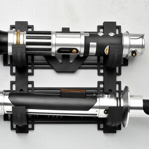May include: A black metal wall mount with two lightsaber holders. The lightsabers are silver, black, and gold.