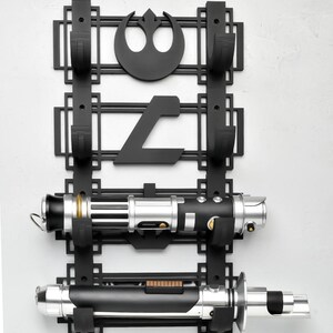 May include: Black wall mount with two hooks for holding lightsabers. The mount features a Rebel Alliance symbol and a symbol from the Star Wars universe. Two lightsabers are displayed on the mount.