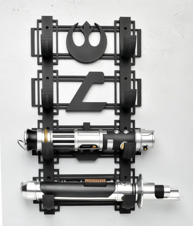 May include: Black wall mount with two hooks for holding lightsabers. The mount features a Rebel Alliance symbol and a symbol from the Star Wars universe. Two lightsabers are displayed on the mount.