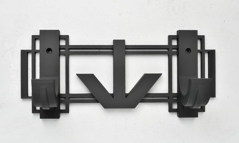 May include: Black metal wall mounted coat rack with an art deco design. The rack has two hooks and a downward pointing arrow in the center.