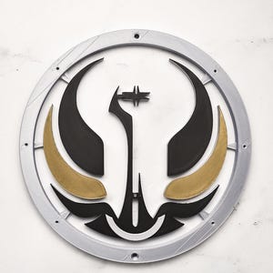 May include: A circular, layered wall art piece with a silver outer ring. The design features black and gold shapes, including curved elements and a central star-like figure. The artwork is set against a white background.