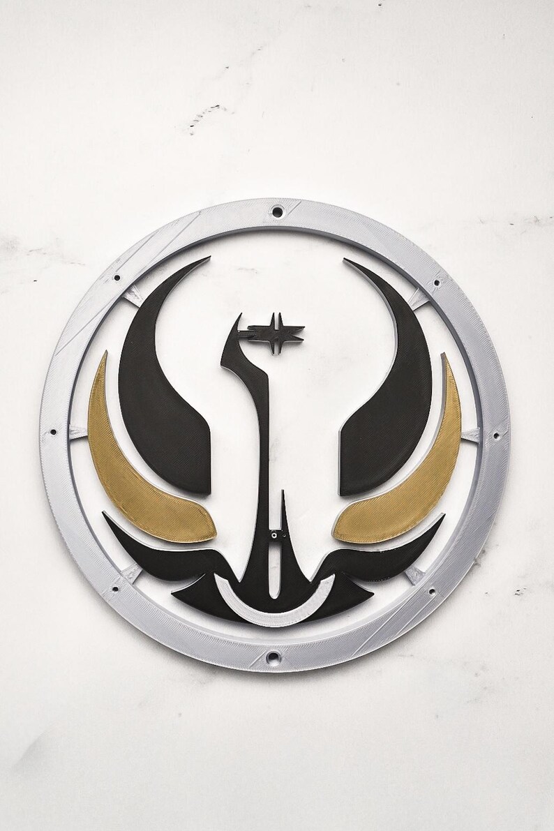 May include: A circular, layered wall art piece with a silver outer ring. The design features black and gold shapes, including curved elements and a central star-like figure. The artwork is set against a white background.