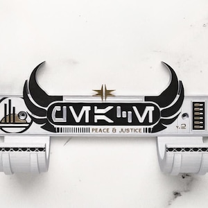 May include: A white and black wall-mounted item with the text "GMKM" and "Peace & Justice". The item has two curved black wings and a gold star. It features two white, curved holders at the bottom.