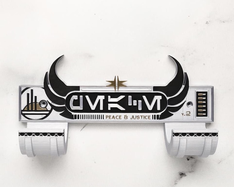 May include: A white and black wall-mounted item with the text "GMKM" and "Peace & Justice". The item has two curved black wings and a gold star. It features two white, curved holders at the bottom.