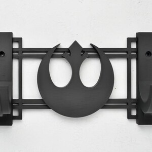 May include: Black metal wall mount with two hooks and a Rebel Alliance symbol in the center. The hooks are designed to hold lightsabers.