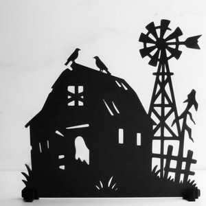 May include: Black metal silhouette of a rural scene featuring a house with two birds perched on the roof, a windmill, and a fence. The design is cut out, creating a negative space effect against a white background.