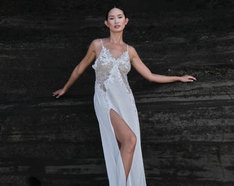 Puede incluir: Un vestido largo de color blanco con un corpiño de encaje transparente con intrincados bordados florales, tirantes finos y una abertura lateral alta que se extiende desde el muslo hasta el dobladillo, creando una silueta fluida.