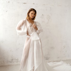 May include: A floor-length white sheer gown featuring voluminous puff sleeves, a V-neckline with delicate lace trim, and a wide fabric sash tied at the waist. The garment is constructed from a translucent, lightweight material with a subtle texture.