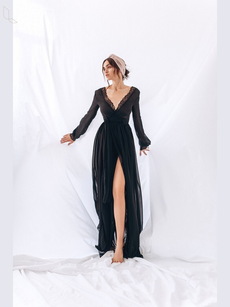 May include: A black maxi dress with a lace trim and a front slit. The dress has long sleeves and a V-neckline. The model is wearing a light pink headband.