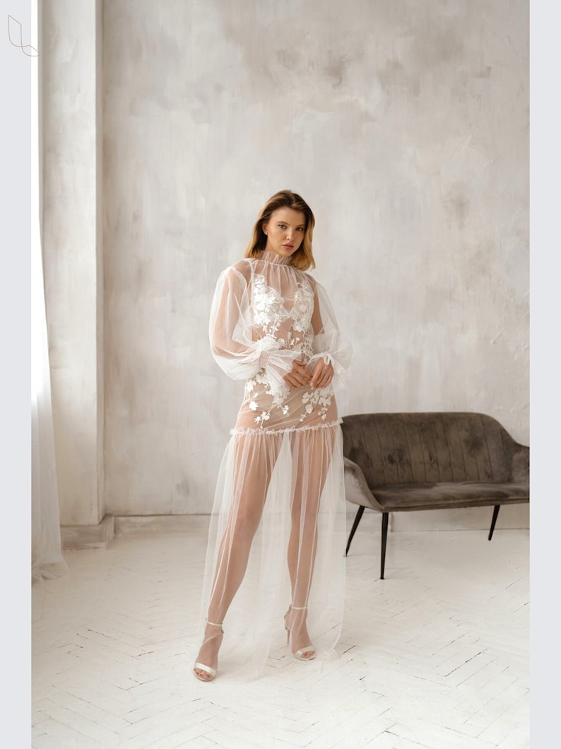Pu&ograve; includere: Un abito maxi bianco in tulle trasparente con ricami floreali e maniche lunghe. L'abito ha un corpetto aderente e una gonna fluida.