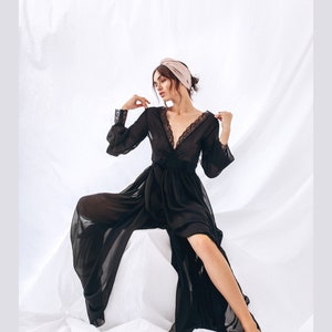 May include: A black sheer robe with lace trim and long sleeves. The robe is worn by a person with a pink turban on their head. The person is sitting on a white surface.