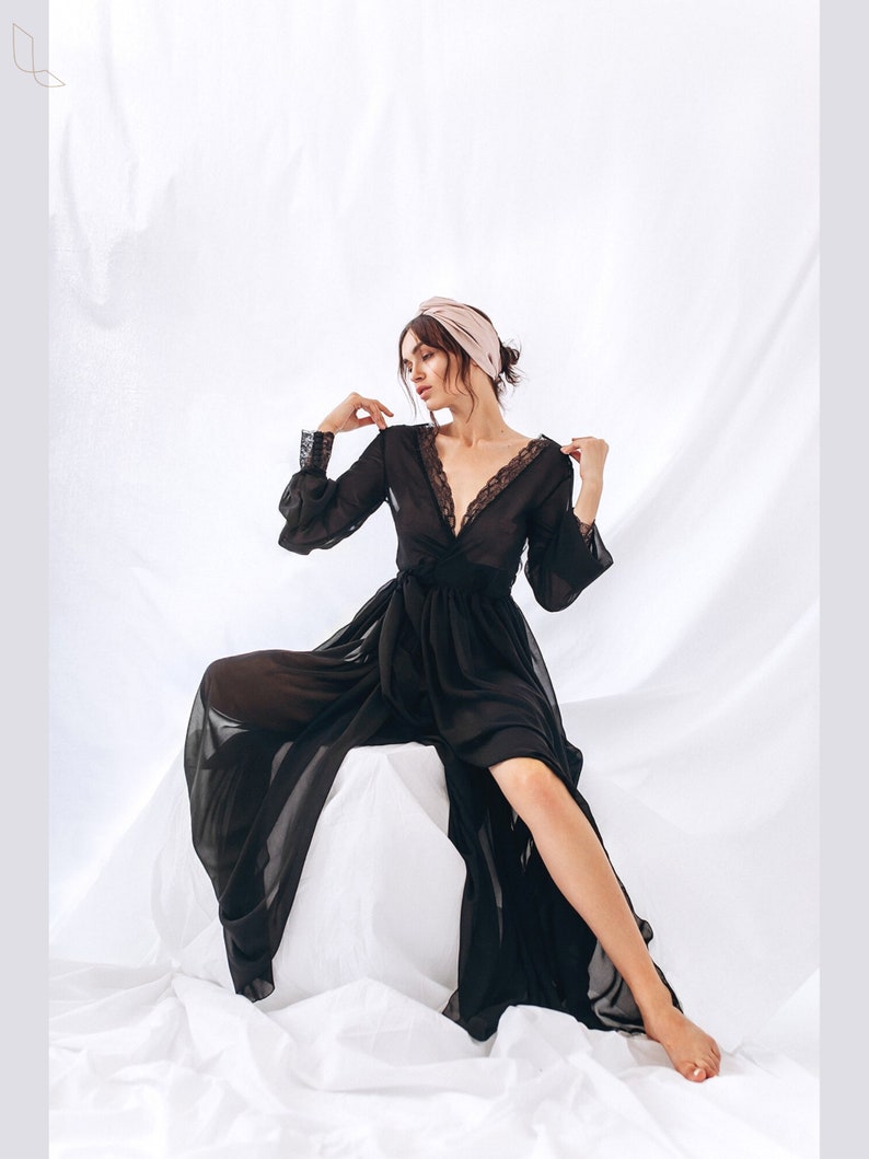 May include: A black sheer robe with lace trim and long sleeves. The robe is worn by a person with a pink turban on their head. The person is sitting on a white surface.
