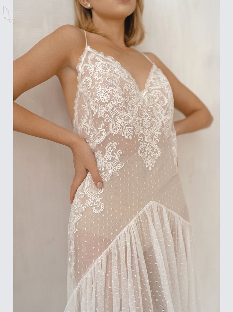 May include: A white lace and tulle dress with a V-neckline and a scalloped hem. The dress has a sheer bodice with delicate floral embroidery and a dotted tulle skirt.