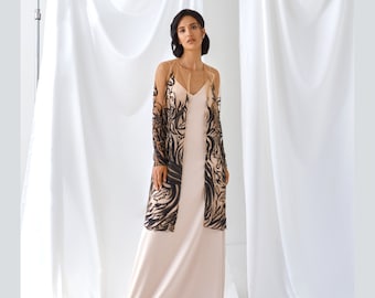 Puede incluir: Una mujer lleva un vestido largo de satén de color nude con una sobrecapa transparente de color negro y nude con un diseño floral. La sobrecapa tiene mangas largas y un escote en V.